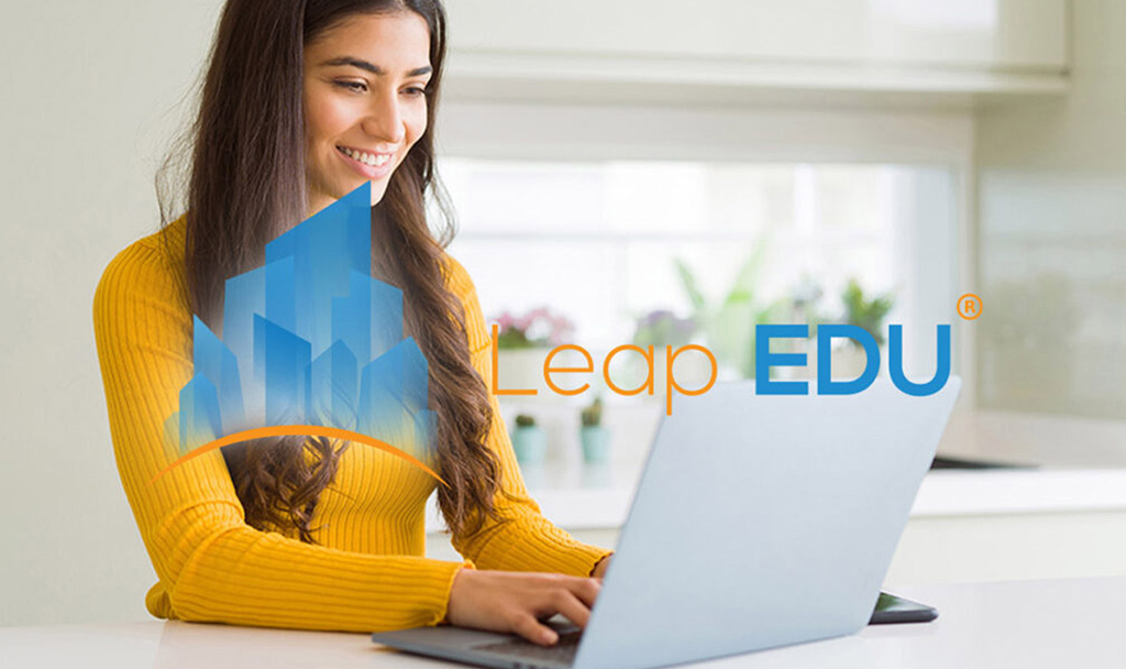 Engaging with Leap EDU Resources | Idea Stream Marketing Woman in a yellow sweater smiling while using a laptop at a bright kitchen table with Leap EDU logo.