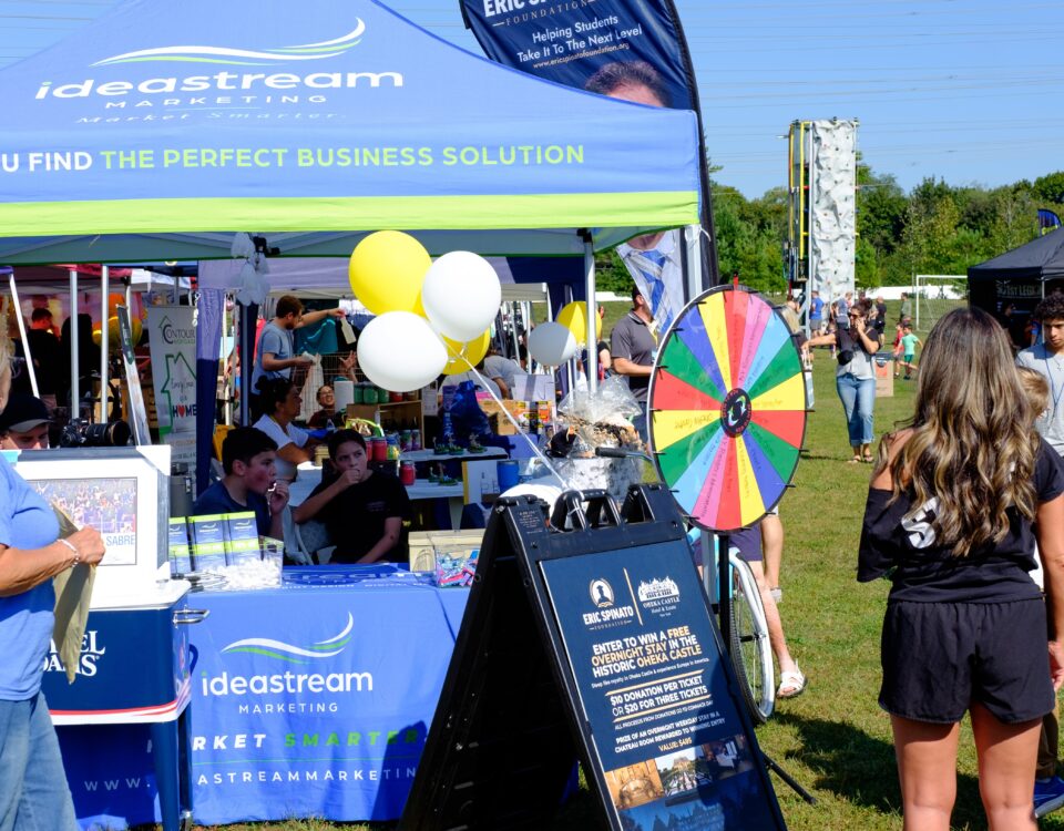Idea Stream Marketing team booth at Commack Day 2025, featuring the prize wheel, community visitors, and local vendors.