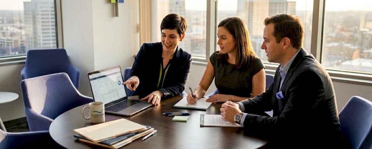 Three professionals in a meeting around a round table in a bright office, discussing a laptop screen with charts as cityscape visible outside the windows, coffee mug nearby.