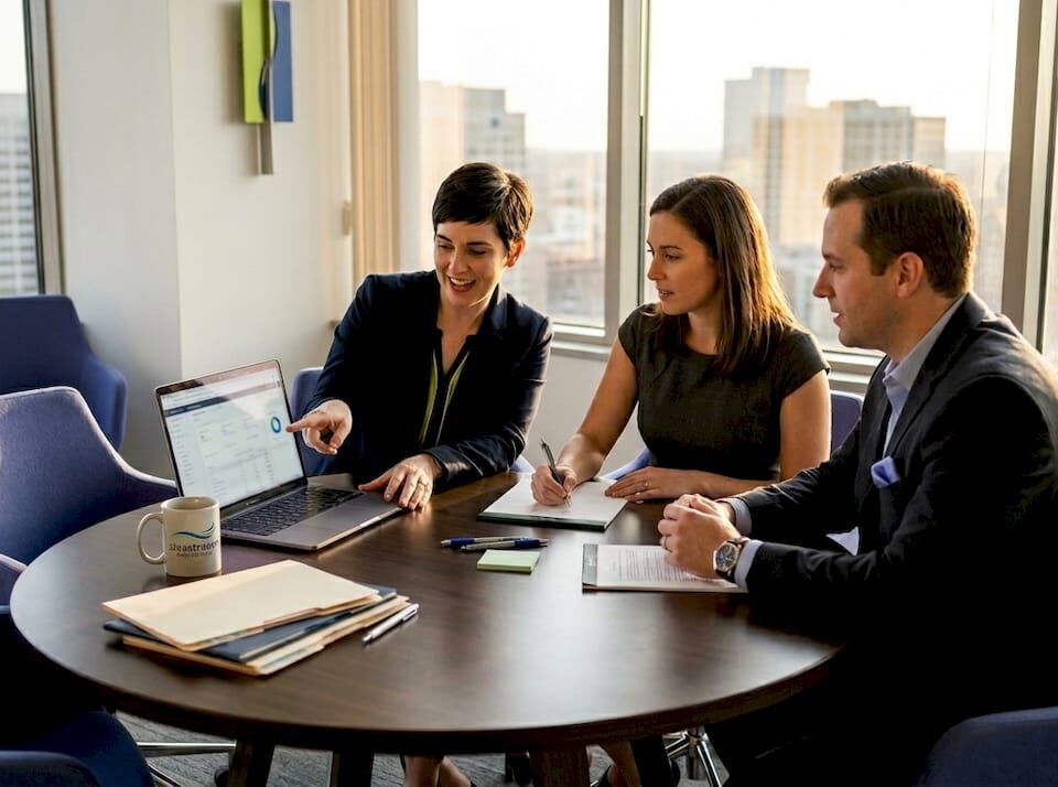 Three professionals in a meeting around a round table in a bright office, discussing a laptop screen with charts as cityscape visible outside the windows, coffee mug nearby.