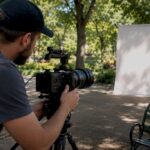 Videographer in a Park Setup | Idea Stream Marketing Videographer setting up a video camera on a tripod in a park with a blank backdrop.