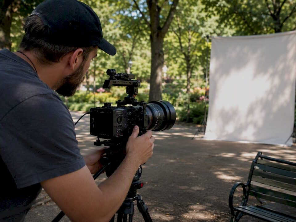 Videographer setting up a video camera on a tripod in a park with a blank backdrop.